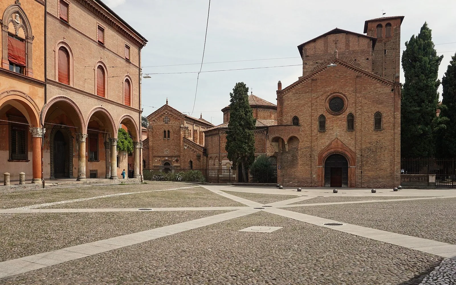 Piazza Santo Stefano a Bologna con il selciato in ciottoli attraversato da fasce di pietra: a sinistra un palazzo con portico ad archi, a destra le facciate in mattoni delle chiese del complesso di Santo Stefano tra alti cipressi