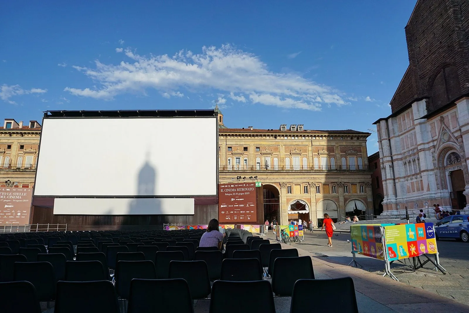 Il grande schermo bianco montato in Piazza Maggiore per Il Cinema Ritrovato con file di sedie nere vuote davanti, di giorno, tra i palazzi della piazza e la facciata della Basilica di San Petronio sotto un cielo azzurro