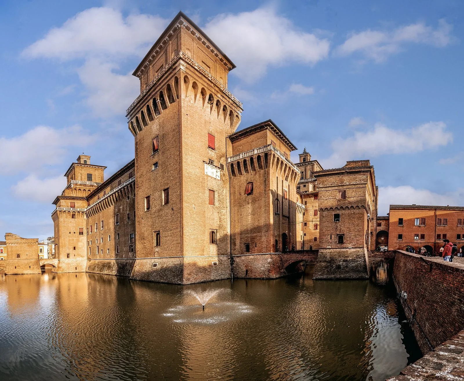 Il Castello Estense di Ferrara con la grande torre in mattoni e i torrioni merlati che si specchiano nell'acqua del fossato, con una piccola fontana al centro e il cielo azzurro con nuvole