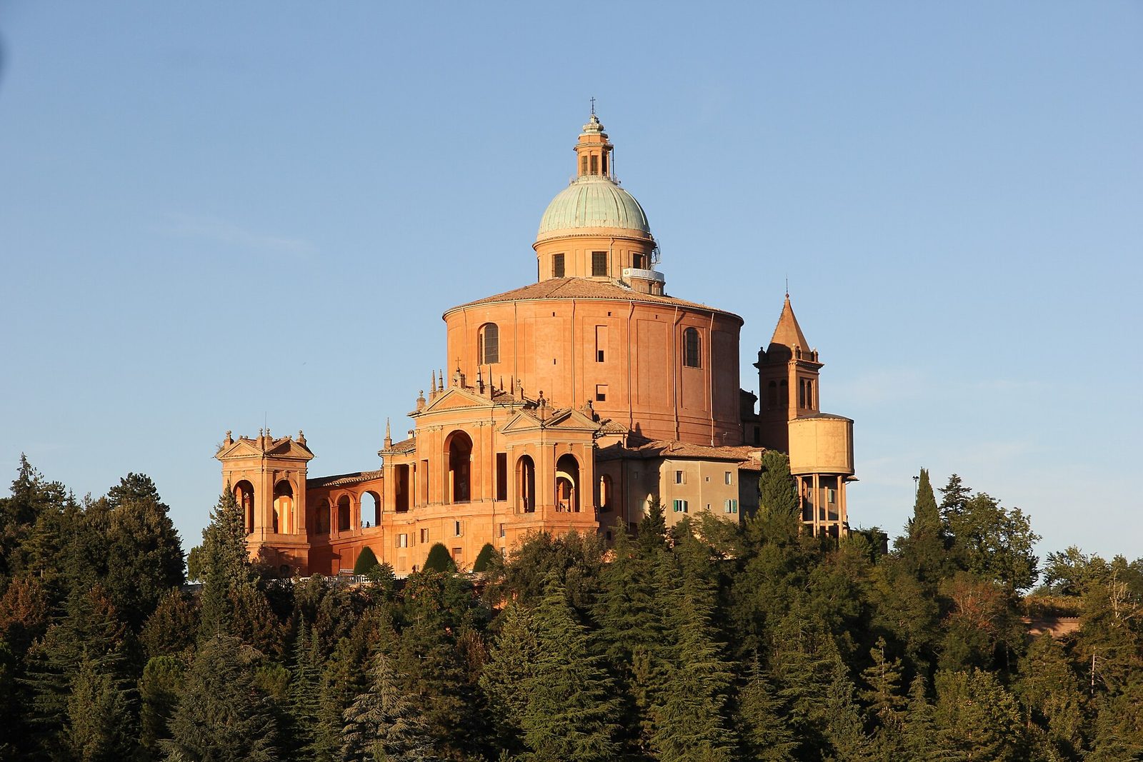Il santuario della Madonna di San Luca in mattoni rosati con la cupola verde, illuminato dalla luce calda del tramonto, che emerge da un bosco di alberi scuri sul colle contro il cielo azzurro