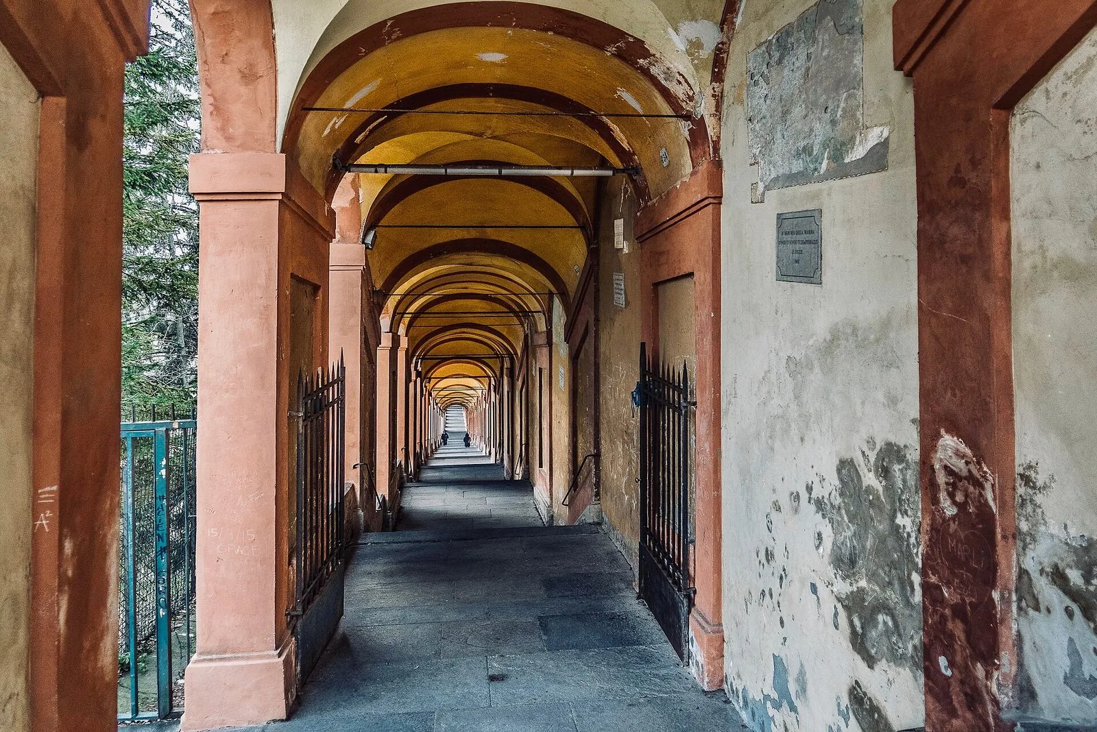 L'interno del portico di San Luca: una lunga fuga di volte a crociera gialle e pilastri rosati che sale con gradini verso il fondo, con due cancelli in ferro aperti in primo piano