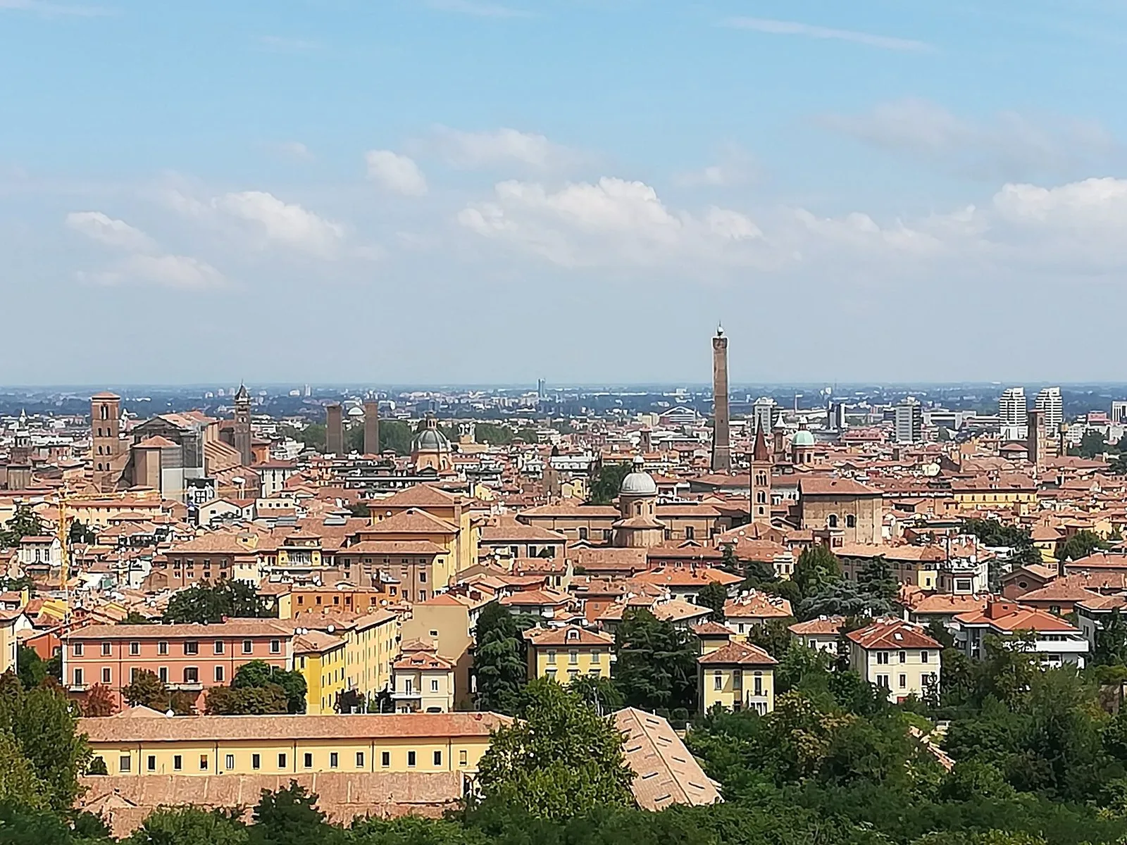 Panorama di Bologna dall'alto dei colli: un mare di tetti rossi e palazzi color ocra, campanili e cupole, con la Torre degli Asinelli che svetta al centro sotto un cielo azzurro